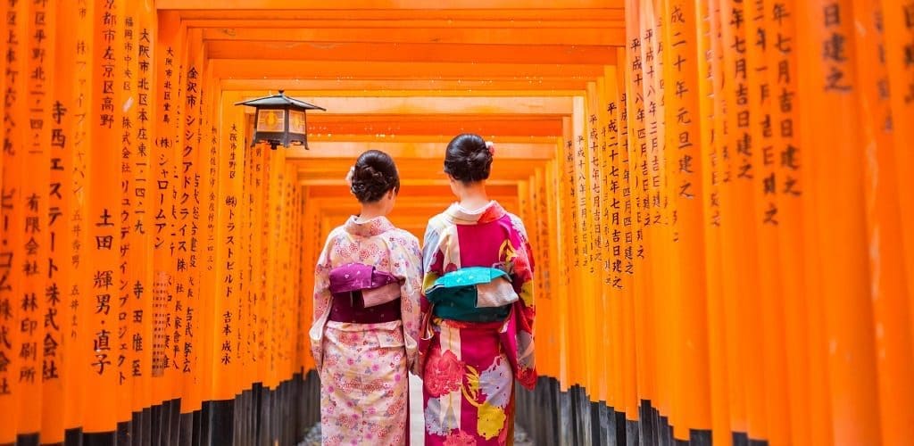 Two geishas among red wooden Tori Gate at Fushimi Inari Shrine in Kyoto, Japan.